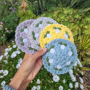 May include: A hand holds a stack of crocheted coasters in various pastel colors: lavender, light green, yellow, and blue. Each coaster features a floral design with white and green accents, perfect for home decor.