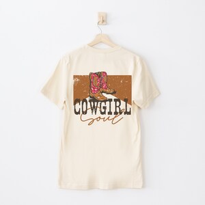 May include: A beige t-shirt with a brown and orange graphic of cowboy boots on the back. The graphic also includes the text "COWGIRL Soul".