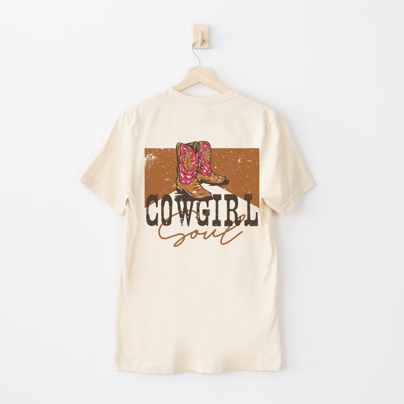 May include: A beige t-shirt with a brown and orange graphic of cowboy boots on the back. The graphic also includes the text "COWGIRL Soul".