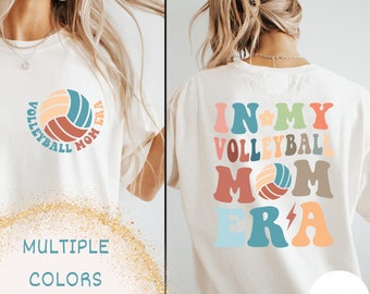 Może przedstawiać: Biała koszulka z napisem "In My Volleyball Mom Era" w kolorowych, retro literach z grafiką piłki siatkowej. Z tyłu koszulki znajduje się grafika piłki siatkowej z napisem "Volleyball Mom Era" w kółku.