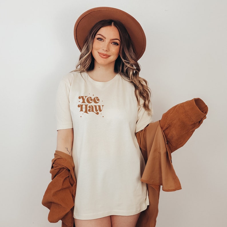 May include: A white t-shirt with a brown graphic that says "Yee Haw" in a retro font. The shirt is being worn by a person with long brown hair and a brown hat.