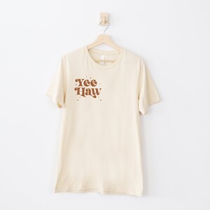 May include: A cream colored t-shirt with a brown graphic that says "Yee Haw" in a retro font.