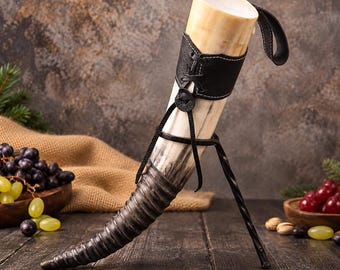 May include: A drinking horn with a natural cream and dark grey gradient finish, featuring a black leather sleeve with white stitching and a lace-up design. The horn is supported by a black metal stand and includes a black leather carrying strap.