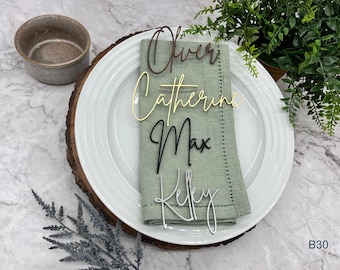 May include: A set of four place card holders with the names Oliver, Catherine, Max, and Kelly written on them. The place card holders are made of wood and are in different colors. The place card holders are on a white plate with a green napkin.