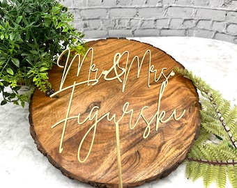 Könnte beinhalten: Goldener Kuchentopper aus Metall mit den Worten "Mr & Mrs [Nachname]" in einer kursiv geschriebenen Schriftart auf einem Kuchenständer aus Holz.