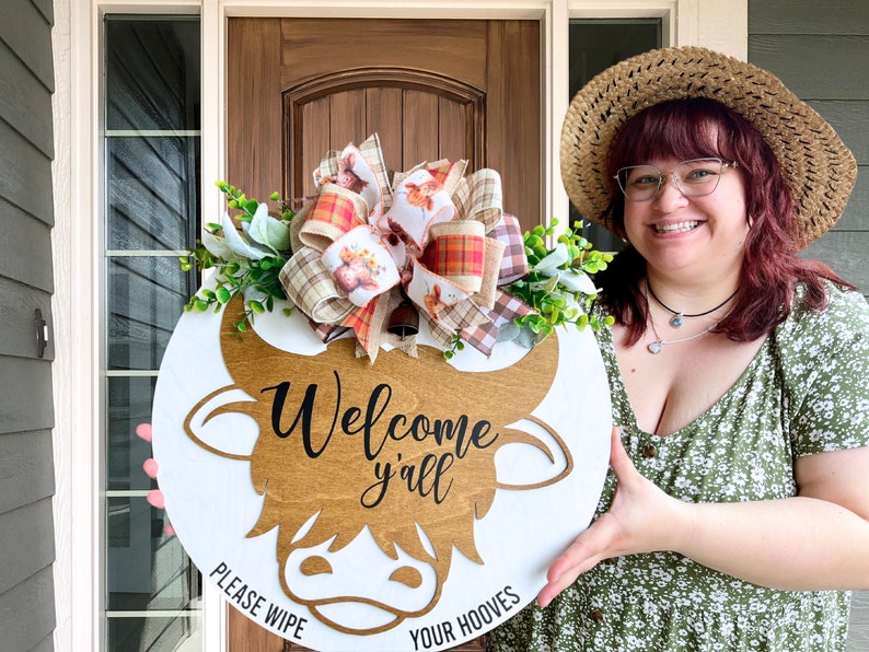 May include: A white wooden doormat with a brown cow head cutout. The cow head has the text "Welcome y'all" and "Please wipe your hooves" written on it. The doormat is decorated with a plaid and floral bow and greenery.