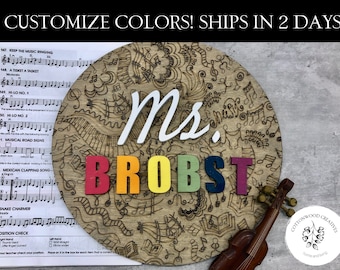 May include: A round wooden sign with a music theme. The sign is decorated with musical notes, treble clefs, and other music-related symbols. The name "Ms. Brobst" is spelled out in colorful letters. The sign is perfect for a music teacher or a music lover.