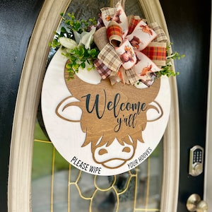 May include: A round wooden sign with a brown cow head cutout. The sign says "Welcome y'all" and "Please wipe your hooves". The sign is decorated with a plaid and floral bow.