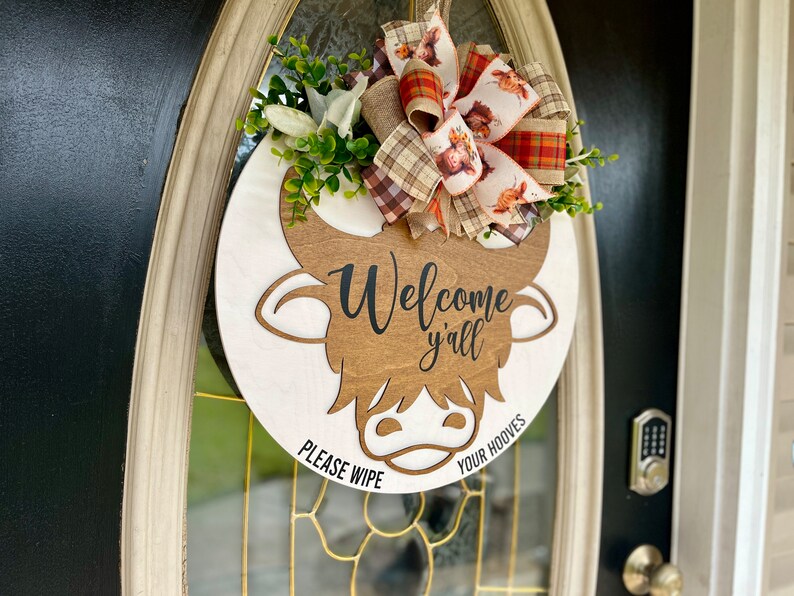 May include: A round wooden sign with a brown cow head cutout. The sign says "Welcome y'all" and "Please wipe your hooves". The sign is decorated with a plaid and floral bow.