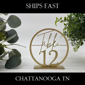 May include: Gold table number sign with the text "Table 12" in a circle. The sign is on a white background.