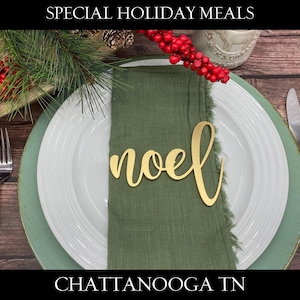 May include: A green napkin with a gold "Noel" place card on a white plate with a green charger. The plate is set on a wooden table with red berries and greenery.