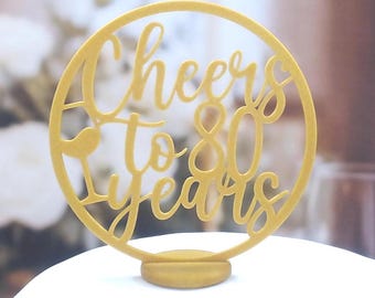 May include: A gold cake topper with the words "Cheers to 80 Years" cut out in a circular design. The topper has a small base for insertion into a cake. The background is blurred, suggesting a celebratory setting.