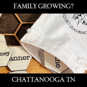 May include: Wooden hexagon name tags with black lettering, scattered on a wooden surface. A canvas bag with a floral logo and the text "Cottonwood Creative" is open, revealing more tags. The text "FAMILY GROWING?" and "CHATTANOOGA TN" are displayed.