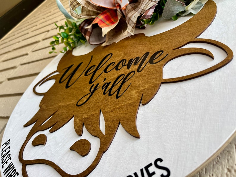 May include: A wooden round sign with a brown cow head cutout. The sign says "Welcome y'all" in black lettering. A fall-themed bow is attached to the top of the sign.
