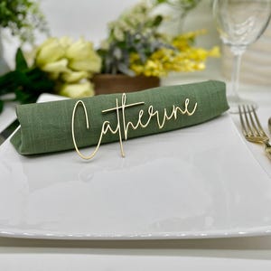 May include: A gold metal name card with the name "Catherine" written in cursive script. The card is placed on a green linen napkin on a white plate.