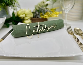May include: A gold metal name card with the name "Catherine" written in cursive script. The card is placed on a green linen napkin on a white plate.