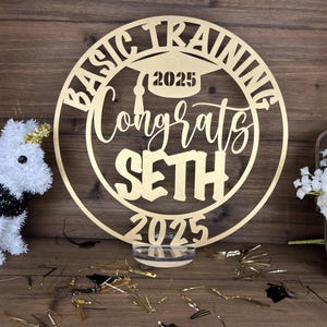 May include: Gold-colored wooden circle with the text "Basic Training Congrats Seth 2025" and a graduation cap with the year 2025 on it. The circle is on a clear acrylic stand.