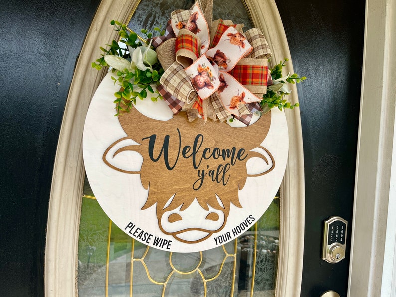 May include: A white and brown wooden door hanger shaped like a cow's head with the text "Welcome y'all" and "Please wipe your hooves". The hanger is decorated with a plaid and floral bow.