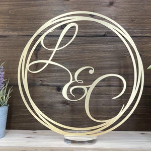 May include: Gold-colored metal monogram sign with the letters "L & C" in a decorative script font. The sign is circular and has a clear acrylic stand.