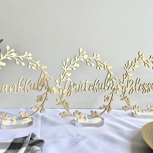May include: Three gold-colored metal wreaths with the words "Thankful", "Grateful", and "Blessed" written in a cursive font. The wreaths are on clear acrylic stands.