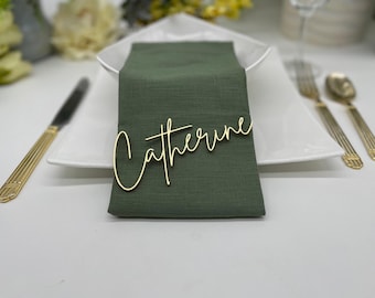 May include: A gold-colored name tag with the name Catherine written in a cursive script font, placed on top of a folded olive green fabric napkin. The napkin rests on a white square plate, accompanied by gold-toned cutlery on the sides.