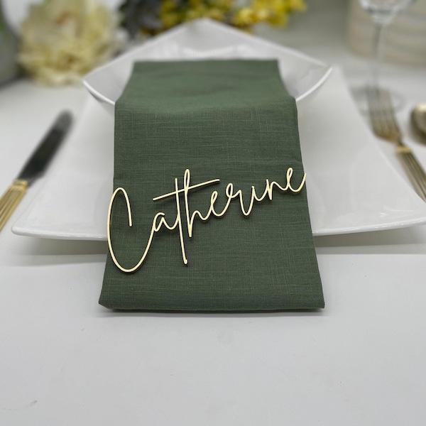 May include: A gold-colored name tag with the name Catherine written in a cursive script font, placed on top of a folded olive green fabric napkin. The napkin rests on a white square plate, accompanied by gold-toned cutlery on the sides.