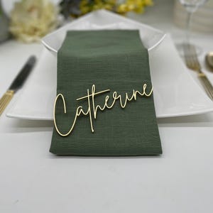 May include: A gold cursive name card with the name "Catherine" on a green linen napkin. The napkin is placed on a white plate with gold silverware.