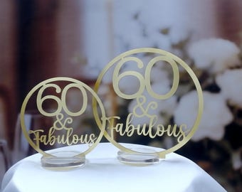 May include: Two gold-colored cake toppers with the text "60 & Fabulous" in a circular design. The toppers are on clear acrylic bases, set against a blurred background of white flowers and glassware. Ideal for a 60th birthday celebration.