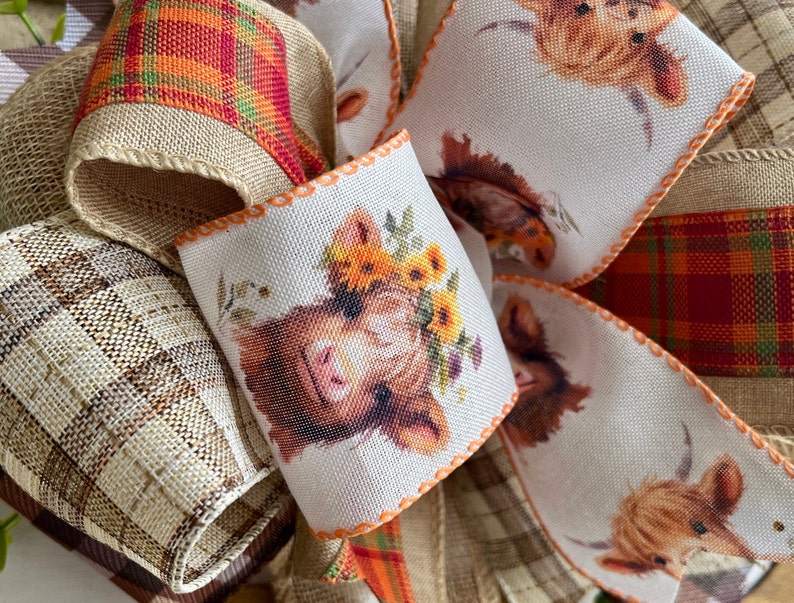 May include: A close-up of a handmade bow with a plaid, burlap, and white fabric with a highland cow print. The bow is decorated with orange and yellow flowers.