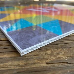 May include: Close-up of the edge of a colorful printed sign with a white corrugated plastic backing.