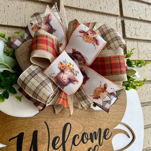 May include: A wooden welcome sign with a brown, tan, and red plaid bow. The bow has a white ribbon with a pattern of highland cows. The sign says "Welcome" in black script.