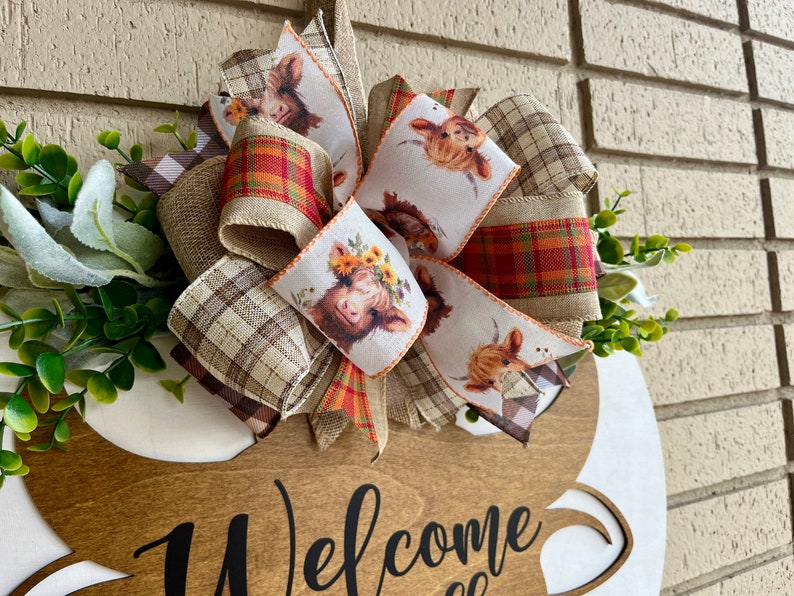 May include: A wooden welcome sign with a brown, tan, and red plaid bow. The bow has a white ribbon with a pattern of highland cows. The sign says "Welcome" in black script.