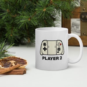 May include: White ceramic mug featuring a video game controller design with the text "PLAYER 2" in black. The controller design is beige with black accents and a heart symbol. The mug is a standard size.