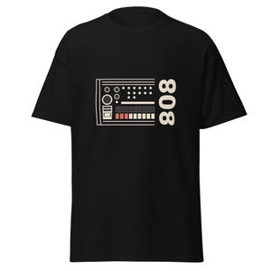 Puede incluir: Camiseta negra con un diseño gráfico crema de una caja de ritmos vintage y el número "808". El diseño incluye círculos, rectángulos y líneas en crema, melocotón y negro. Una camiseta unisex informal.