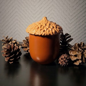 Peut inclure: Une tasse en céramique de couleur marron en forme de gland avec un dessus texturé. La tasse est entourée de pommes de pin sur une surface noire.