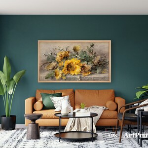 May include: A framed floral print featuring a bouquet of sunflowers in shades of yellow and green. The artwork hangs above a brown leather sofa in a living room setting. The room has a teal wall and a patterned rug.