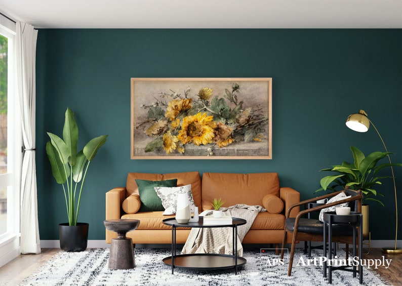May include: A framed floral print featuring a bouquet of sunflowers in shades of yellow and green. The artwork hangs above a brown leather sofa in a living room setting. The room has a teal wall and a patterned rug.