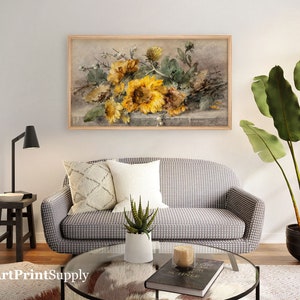 May include: A framed floral print featuring sunflowers in shades of yellow and green. The artwork hangs above a gray and white patterned sofa with decorative pillows. A black lamp and side table are to the left, and a large potted plant is to the right.