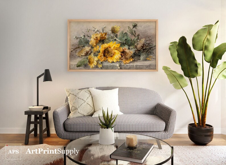 May include: A framed floral print featuring sunflowers in shades of yellow and green. The artwork hangs above a gray and white patterned sofa with decorative pillows. A black lamp and side table are to the left, and a large potted plant is to the right.