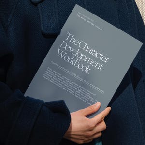 May include: A person holding a gray workbook titled "The Character Development Workbook." The book has white text and is held against a dark navy blue coat. The person is wearing a light blue sweater underneath the coat.