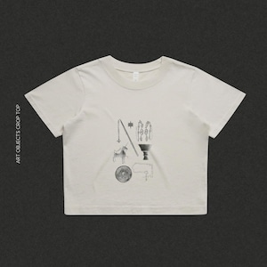 Puede incluir: Top corto color crema con cuello redondo y mangas cortas. La camiseta presenta un diseño gráfico en blanco y negro de varios objetos, incluyendo una cabra, una estrella y una luna. El texto "ART OBJECTS CROP TOP" está impreso en el lado izquierdo y "SAINT VIOLET CREATIVE" en la parte inferior.