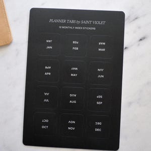 May include: Black rectangular planner tabs by Saint Violet with 12 monthly index stickers. Each tab is labeled with a month in white text. The months are Jan, Feb, Mar, Apr, May, Jun, Jul, Aug, Sep, Oct, Nov, and Dec.