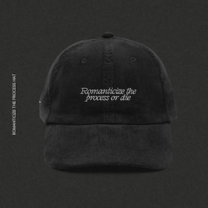 Peut inclure: Casquette de baseball noire en velours côtelé avec le texte brodé "Romanticize the process or die". La casquette a une visière incurvée et un petit bouton centré sur le dessus. Le texte "SAINT VIOLET CREATIVE" est en bas.