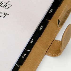 May include: Close-up of a brown leather bound planner with black tabs labeled with the months of the year: January, February, March, April, May, June.