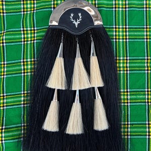 Könnte beinhalten: Schwarz-weißer Kilt-Sporran mit einer silbernen Schnalle und einem Hirschkopf-Design. Der Sporran hat vier weiße Quasten.