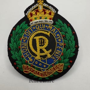 May include: A black embroidered patch with a gold crown, a gold 'R' with 'III' inside, and the text 'HONI SOIT QUI MAL Y PENSE' around the 'R'. The text 'ROYAL ENGINEERS' is on a blue and gold ribbon below the 'R'. The patch is surrounded by a green wreath with red berries.
