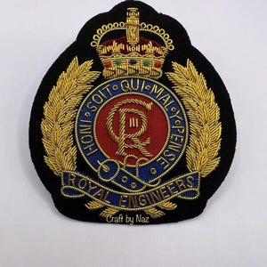 Può includere: Toppa ricamata dei Royal Engineers con sfondo nero. Presenta una corona dorata, foglie di alloro dorate e un emblema centrale con dettagli rossi e blu. Il testo "Royal Engineers" è visibile.