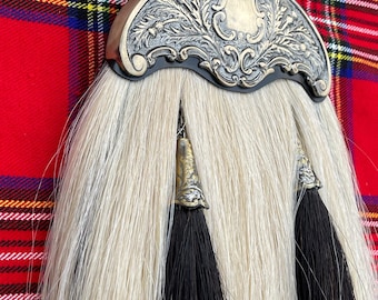 Puede incluir: Un sporran escocés tradicional con un cantle de metal decorativo con un diseño ornamentado. El sporran tiene un cuerpo de piel blanca con dos borlas negras. El fondo es un patrón de tartán rojo y negro.
