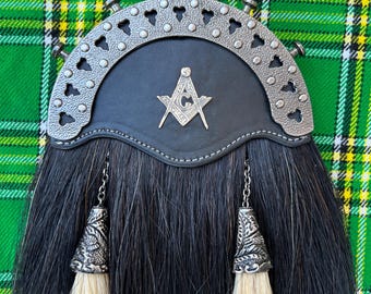 Puede incluir: Sporran de kilt de cuero negro con una hebilla plateada que presenta un símbolo masónico. El sporran tiene dos borlas de crin de caballo blanco con adornos plateados.