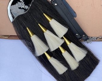Puede incluir: Un sporran negro con un cantle plateado con un emblema de ciervo. El sporran tiene un cuerpo de piel negra, seis borlas blancas con conos de metal dorado y una correa de cuero negro con una cadena.